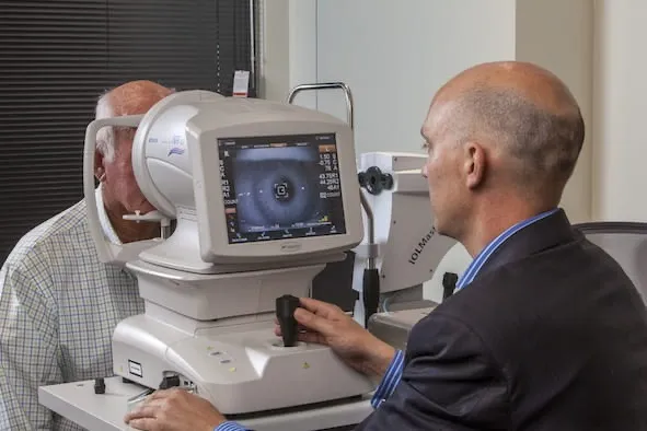 Paciente idoso com o queixo apoiado num aparelho de biometria óptica, enquanto o médico, sentado do outro lado, acompanha a imagem do olho na tela e ajusta o aparelho.