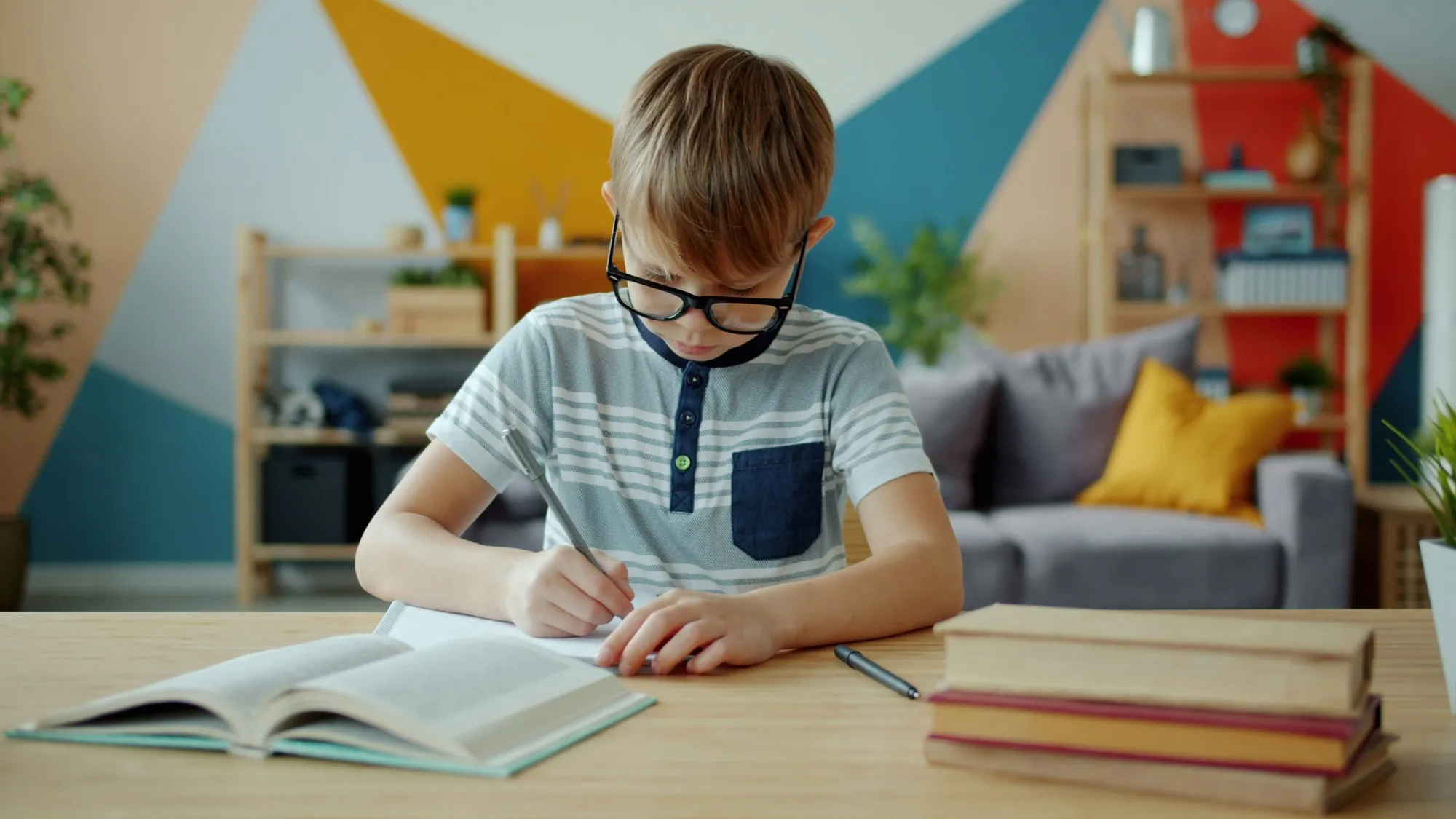 Menino de óculos escrevendo num caderno, sentado à mesa de casa com livros abertos.