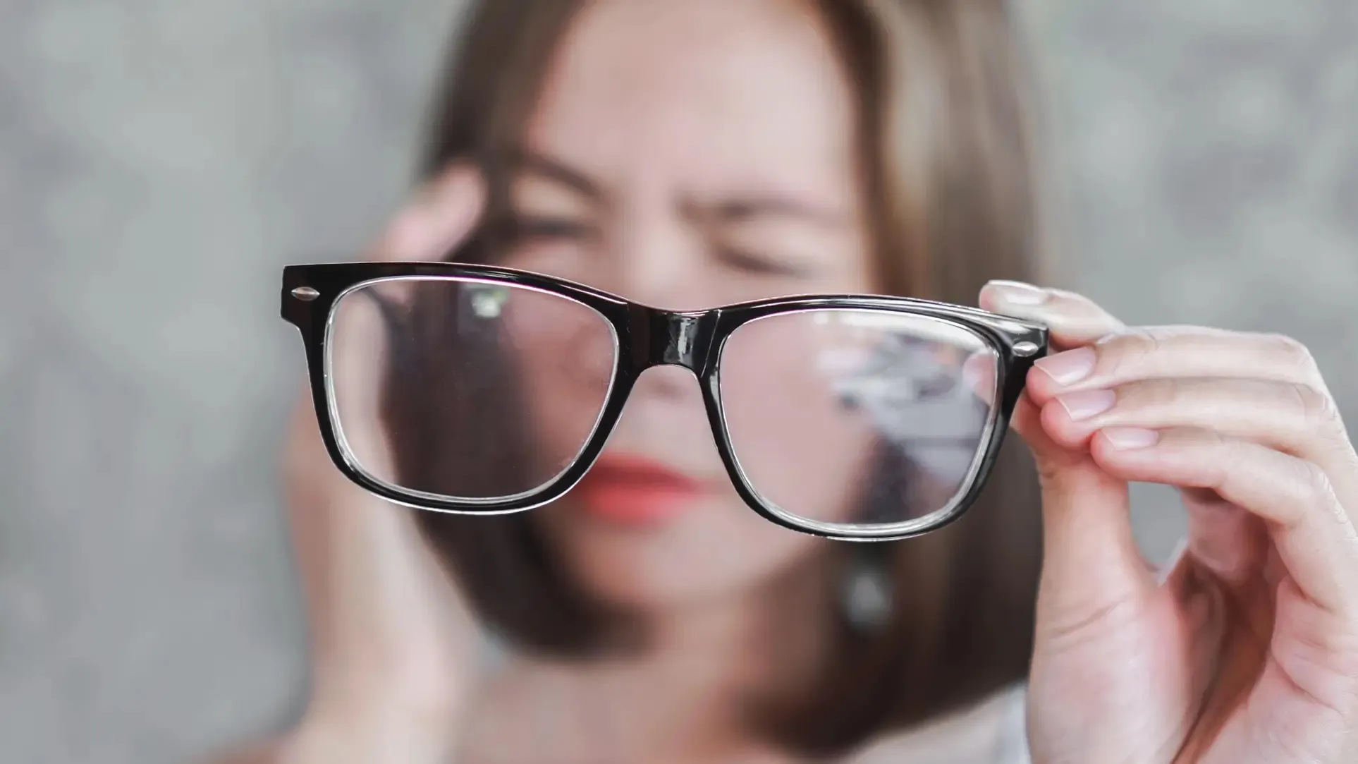 Mulher adulta, desfocada ao fundo, aperta os olhos e leva a mão à têmpora enquanto segura um par de óculos à frente do rosto.